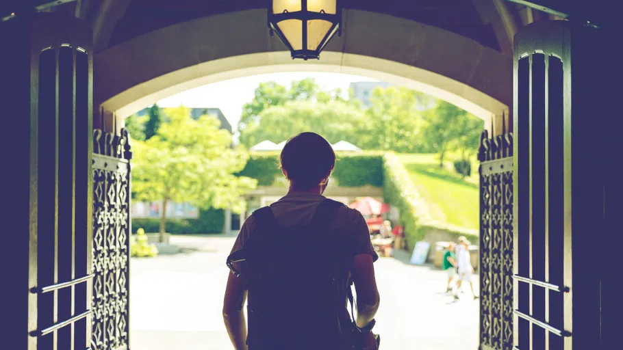 Seen from behind, a student in silhouette exits a building through an iron gate onto a brightly lit outdoor plaza. 