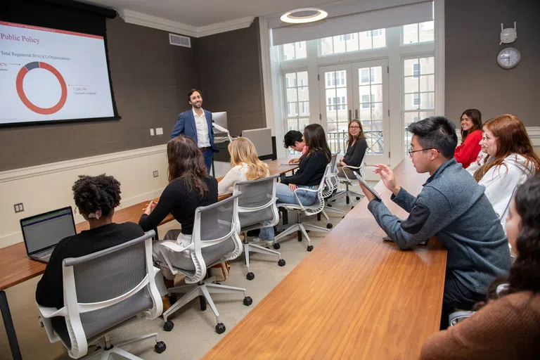 A professor talks with a group of students, who are seated at two long lecture tables. Behind him is a projector screen; the headline “Public Policy” is visible. 