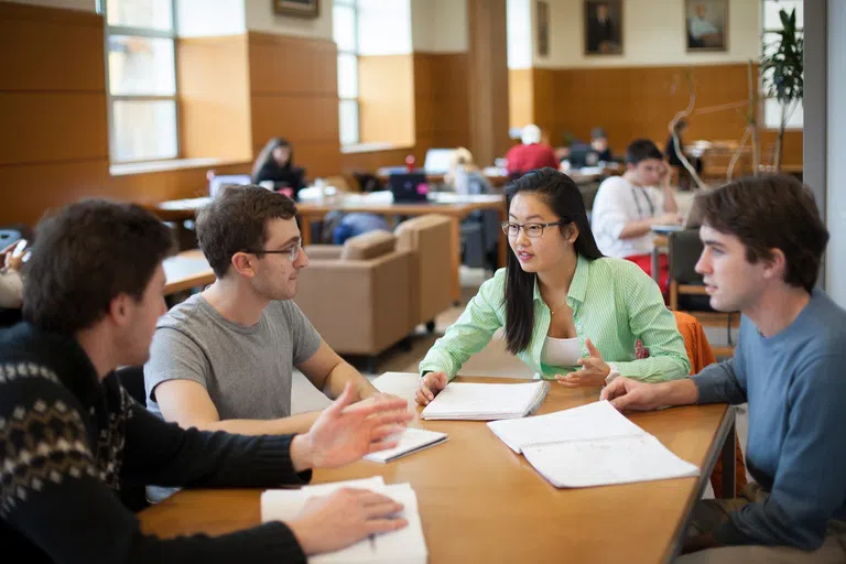 Four students discuss a project at a library table. Each student has a small pile of papers on the table in front of them.