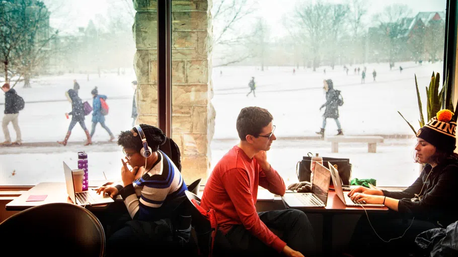 Two students eat at a booth. Behind them is a large plate-glass window looking out onto an academic quad covered in snow. 