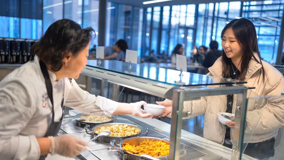 A Cornell Dining staff member hands a bowl to a customer. Between three are three large containers of buffet-style food.