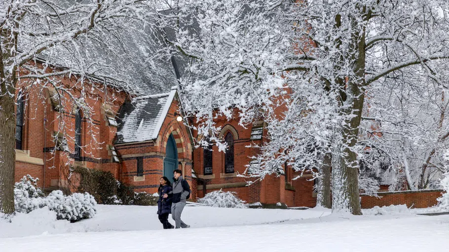 Two students pass by the chapel on a winter day. A layer of snow covers the ground and the tree in front of the building.