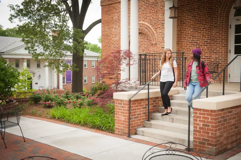 Two wowen with backpacks, one in a white shirt and one in a red jacket, speak to one another on the front stairs of a covered porch at the entrance to a brick building.