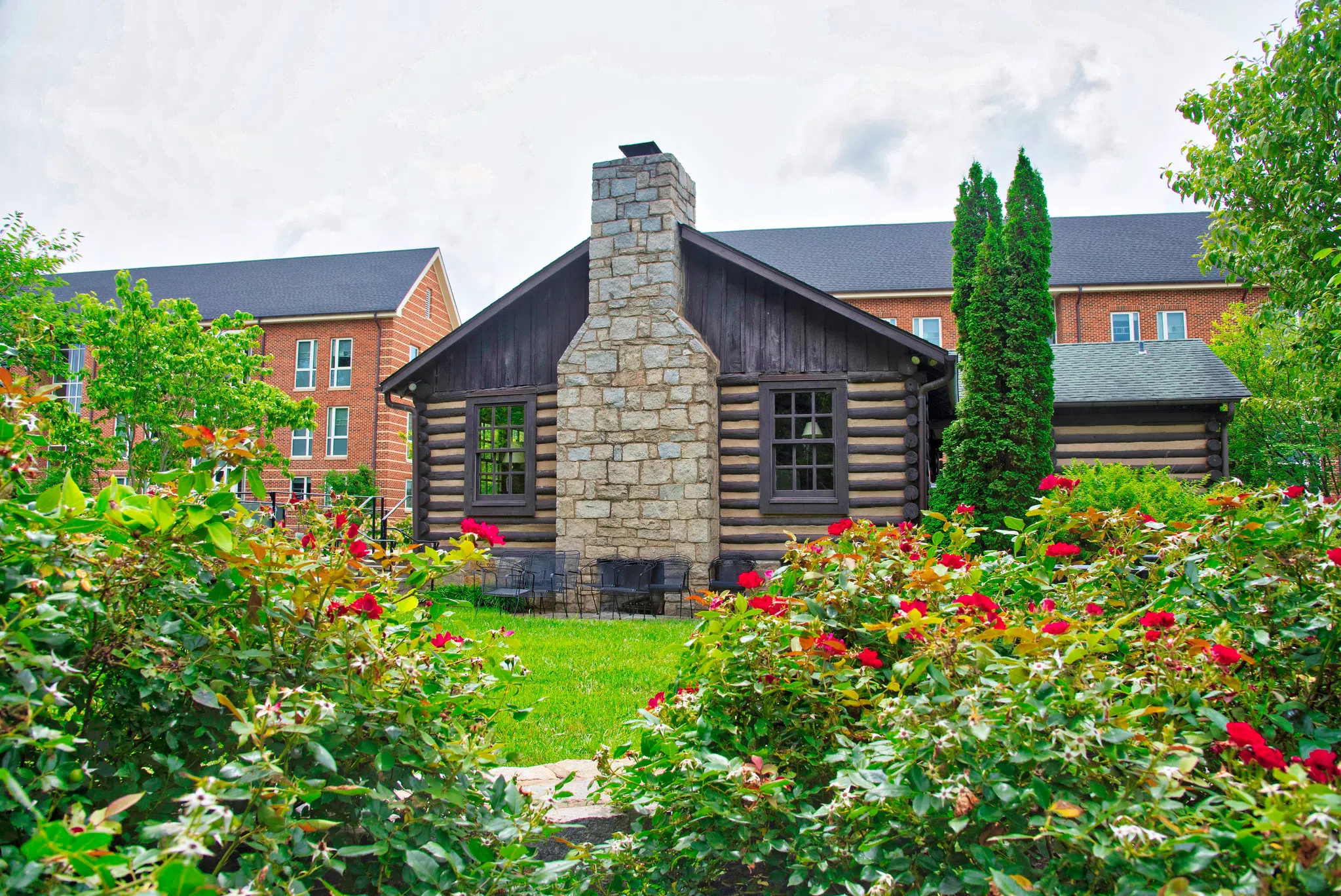 A stone fireplace extends out between two windows in an exterior view of a log cabin. Two three-story brick building can be seen behind the cabin..