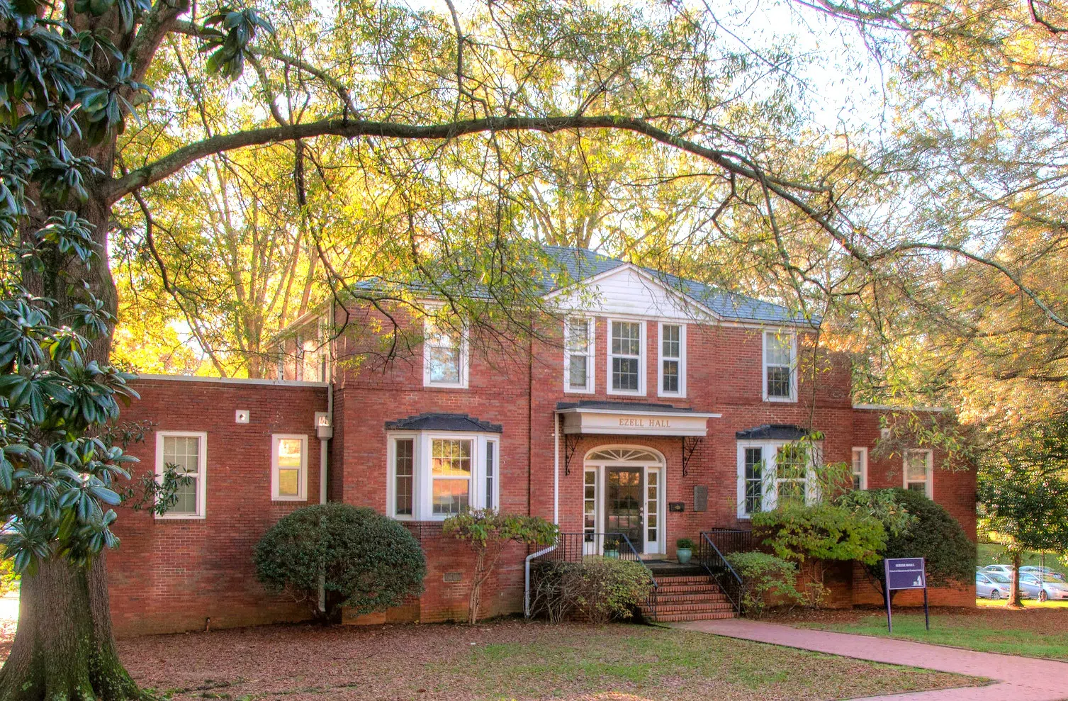 Angled view from left of brick two-story building with green-space and tree in foreground.