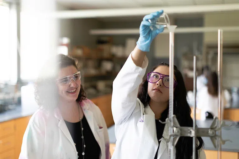 A student in a while lab coat looks reaches up to pour something from a small beaker into the top of a tall, skinny funne.