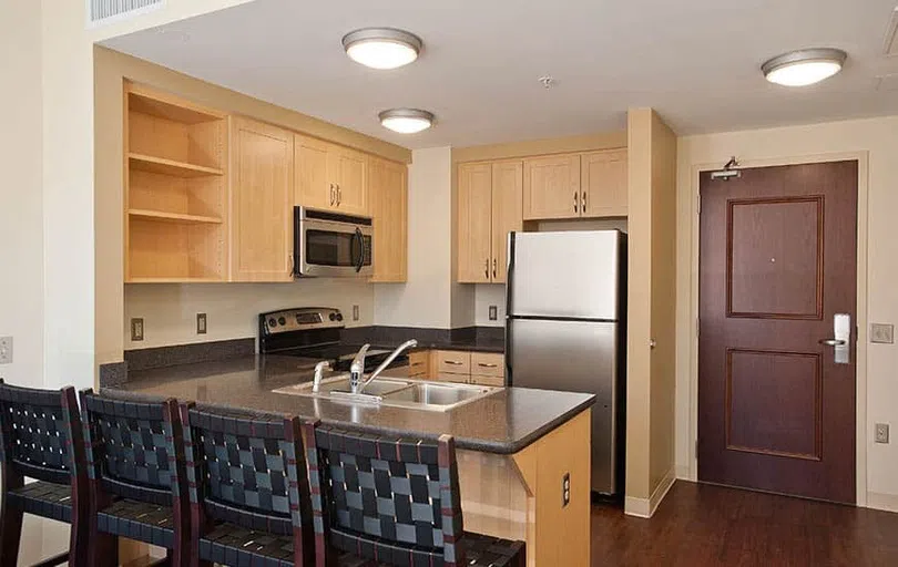 A kitchen with stainless steel appliances and blonde wood cabinets. Four bar stools sit against a counter overlooking the kitchen.