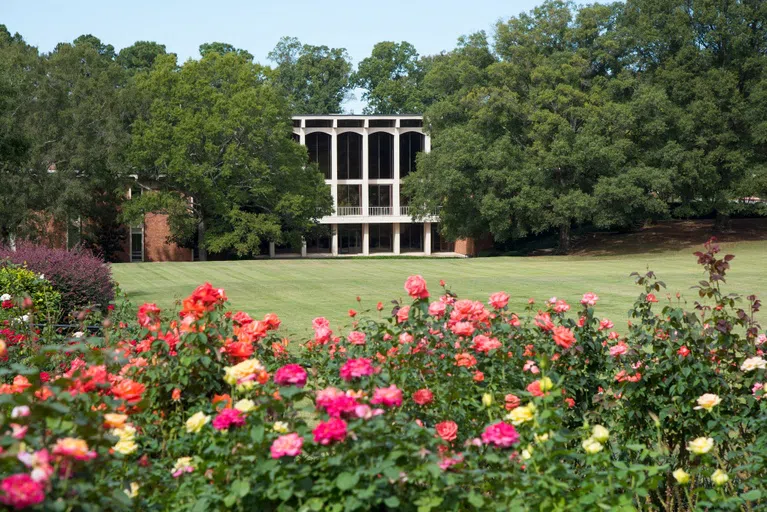 A two-story brick building with tall, arched windows sits in the distance across a grassy lawn flanked by huge trees; yellow, pink and red rosebushes are in the foreground.