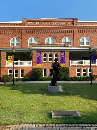  Three story brick building with arched windows. Words at the top of the building read: Zimmerli Performance Center. Words above the center windows of the second story read: Twichell Auditorium.Two purple banners frame the center windows and read: Converse University 1898. In the foreground, a bronze sculpture of a woman singing appears in a grassy square and is flanked by two bronze lampposts adorned with small banners. One banner reads: We will together. The other reads Converse 1889.