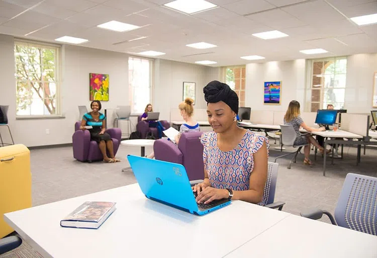 Students in a brightly lit room work with computers at desks or in chairs.