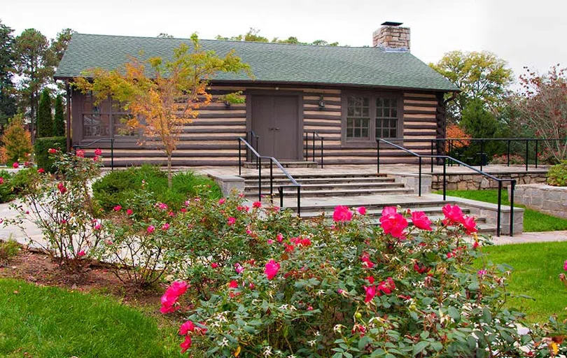 Log cabin building green roof appears behind bright pink flowers in the foreground.