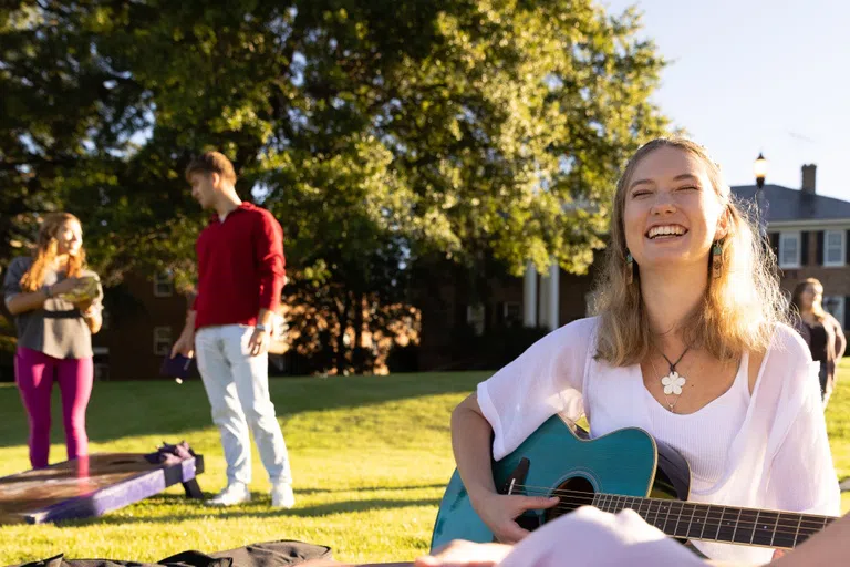 Girl playing guitar outdoors with female listener in the foreground and four people playing games in the background.