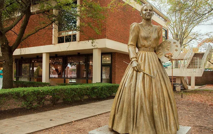 A large bronze statue stands in the foreground shadowed by a two-story building fronted with green shrubs.