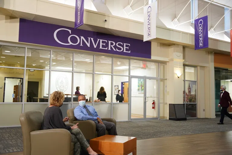 A Converse banner hangs above an interior entrance behind a wall of windows. Students sit in lobby chairs in the foreground.