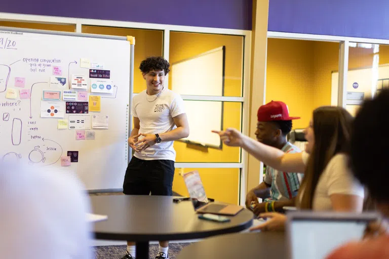 A student stands in front of a large whiteboard while peers in the foreground appear to comment on his work.