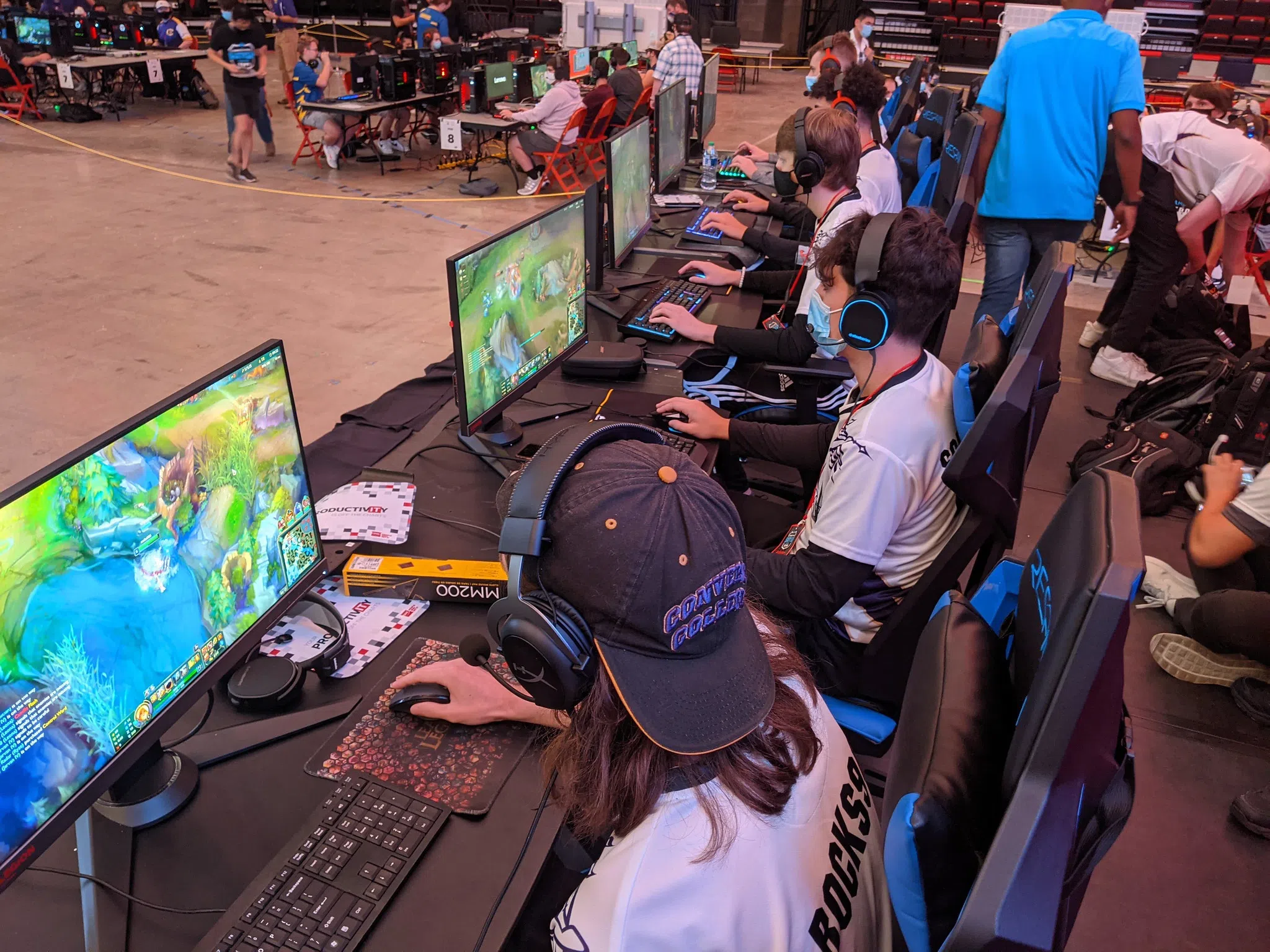 Five students sit at computer stations wearing headphones in an arena with other grouped stations of students.