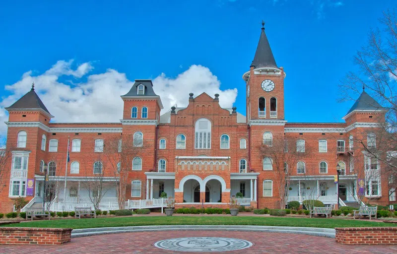 A three-story brick building with arched portico and two towers, one featuring a clock, is shown against a bright blue sky.