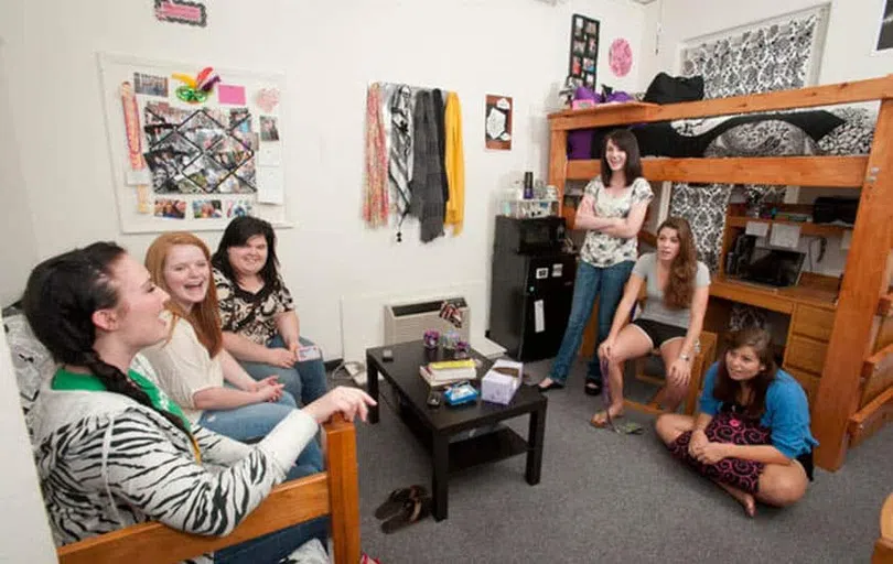 Six female students talk together in a dorm room. Three sit on a sofa and three lean against wooden bunk beds. A rack with scarves hangs on the wall in the center.