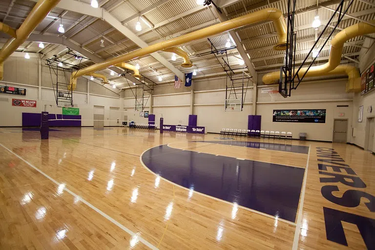 A large empty gymnasium with wood floors accented with purple zones. Yellow piping spans the ceiling.