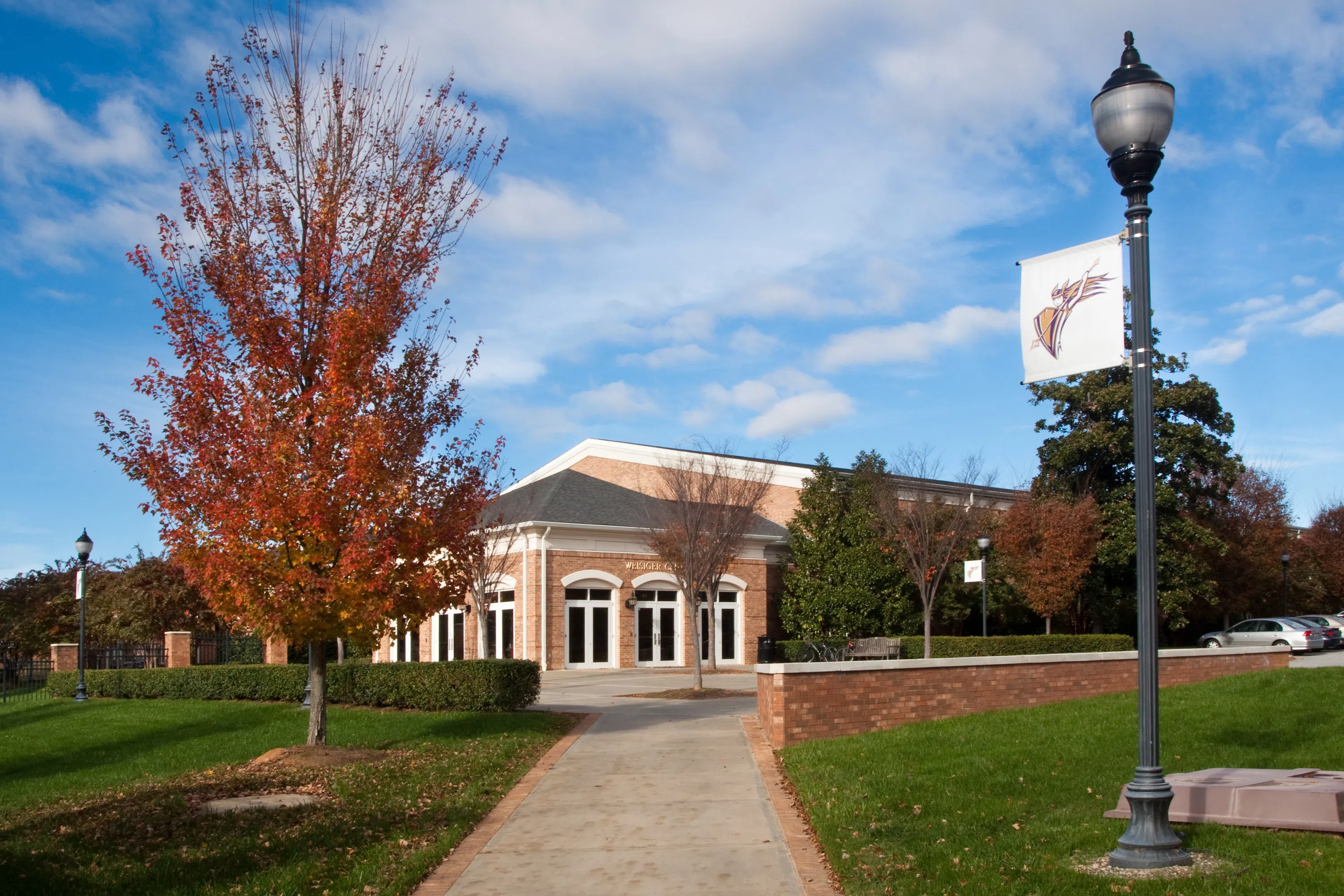 A brick building sits in the distance beyond a pathway flanked by fall trees. A blue cloud-filled sky is behind it.