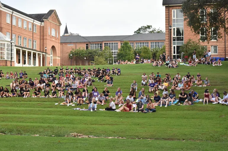 Dozens of students sit on a grass slope with a U-shape of brick buildings in the background.