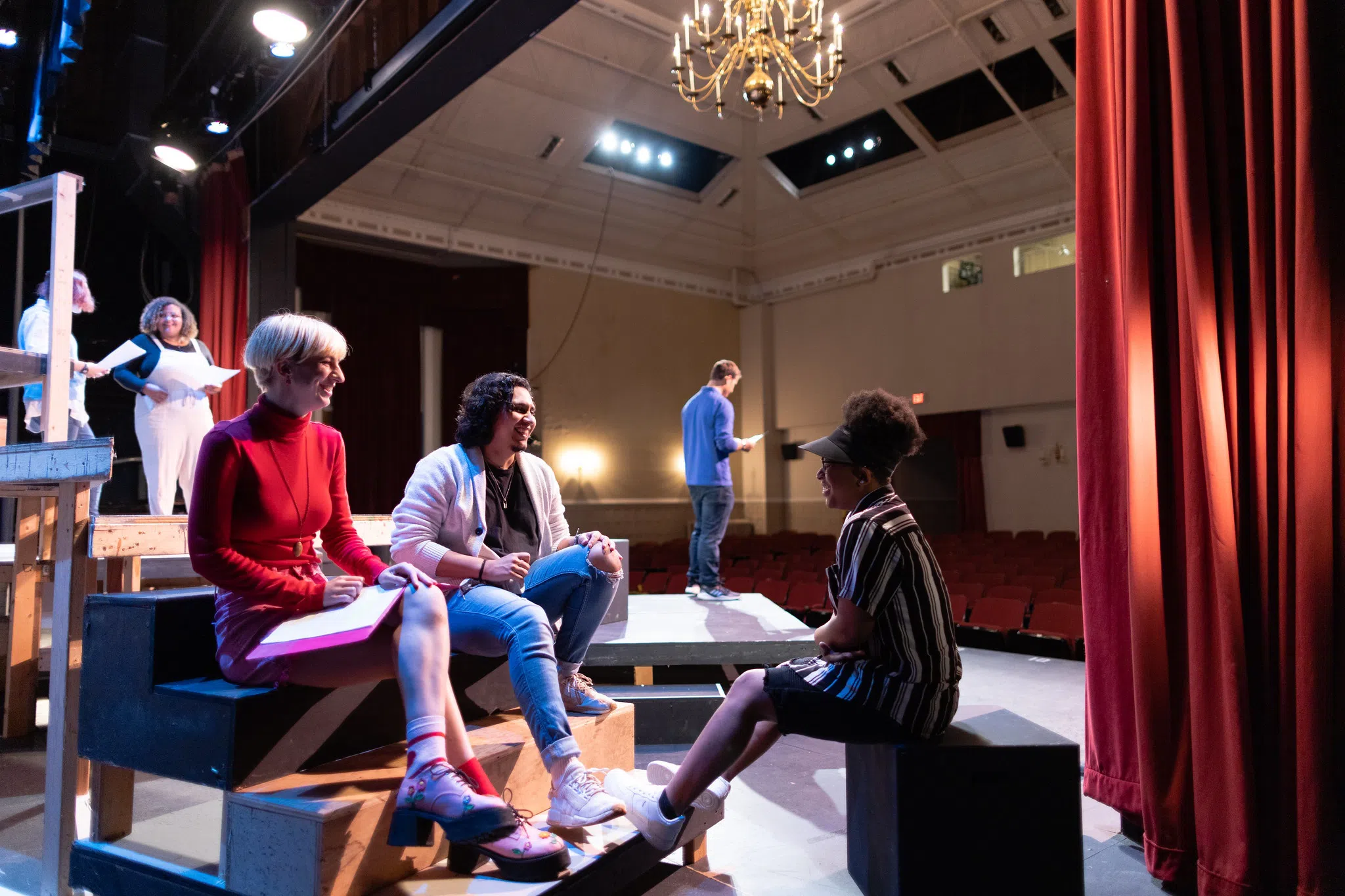 Three students sit on small risers in the foreground and a student studies lines standing in the background. Theatre seats are in the distance.