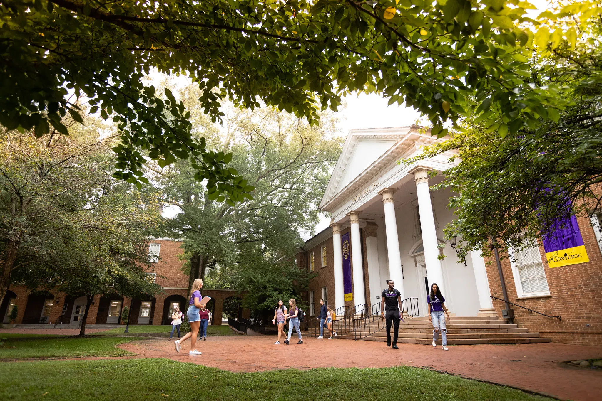 Students walk on brick walkways in front of a two story brick building featuring a four-column portico painted white.