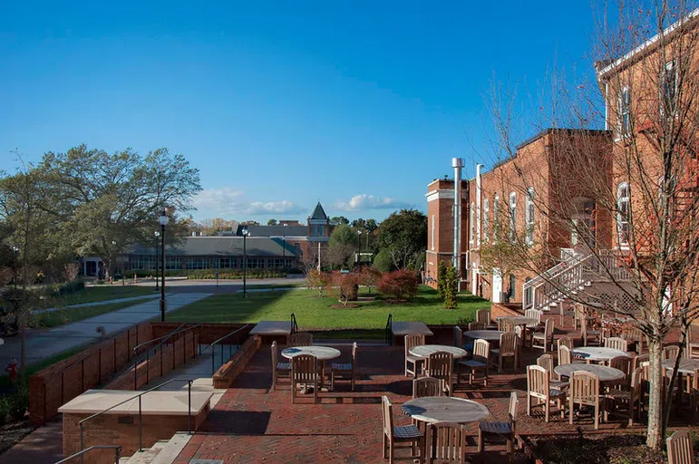 Photo of outdoor, bricked patio on sunny day. Brick buildings, wooden picnic tables and chairs are shown.