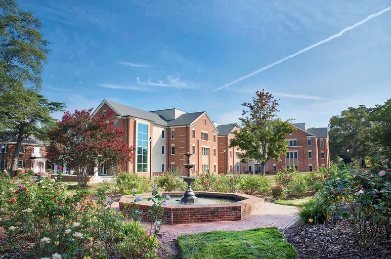 A three-story brick building sits in the distance past a fountain and colorful rose garden.