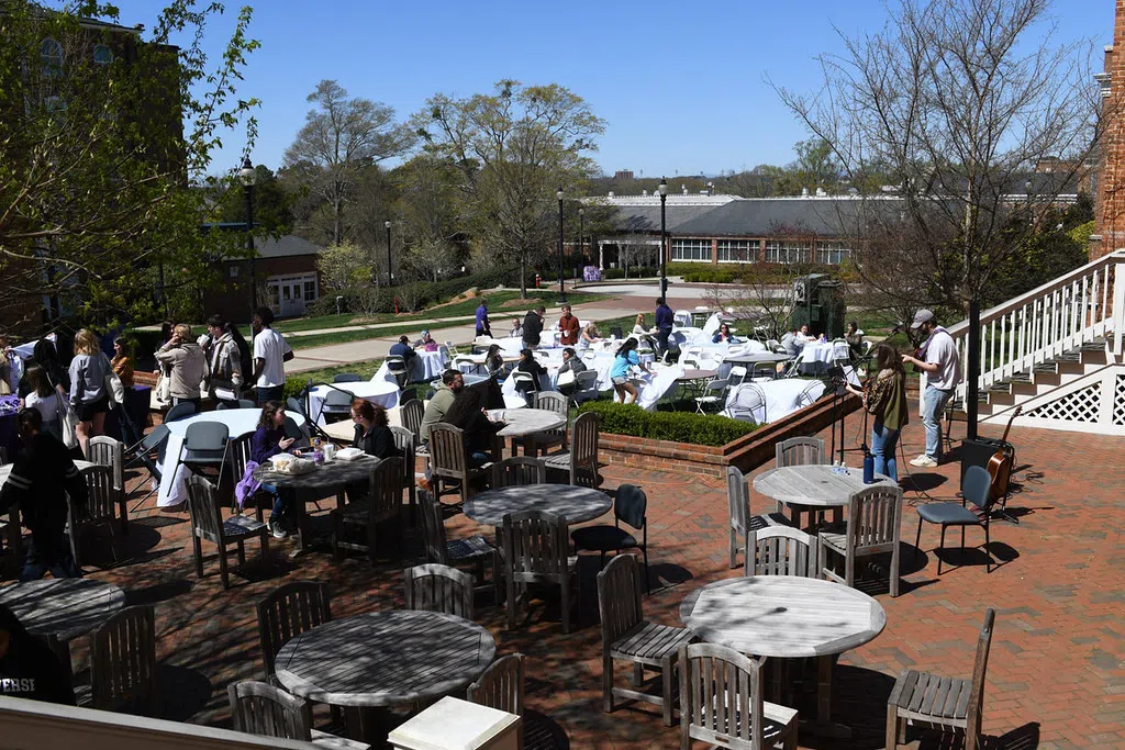 Students and faculty sit at a wooden outdoor table on a sunny day. Another table with students as well as a two story brick building appear behind them.