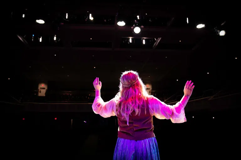 A woman with blonde hair is shown from behind with the audience looking on. Her hands are raised in the air.