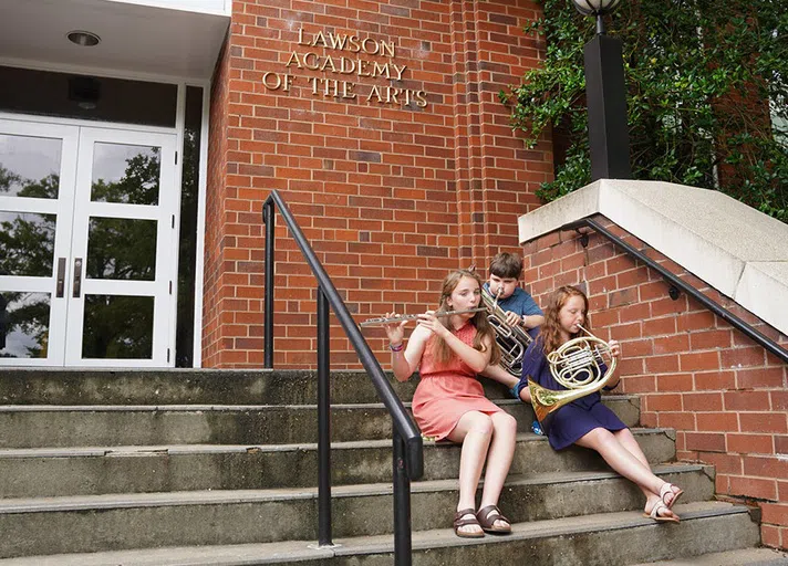 Three children play wind instruments outdoors on the stairs of a brick building. 