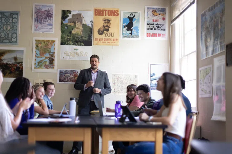 An instructor faces the camera and a table of students with a variety of posters hanging on the wall behind him.