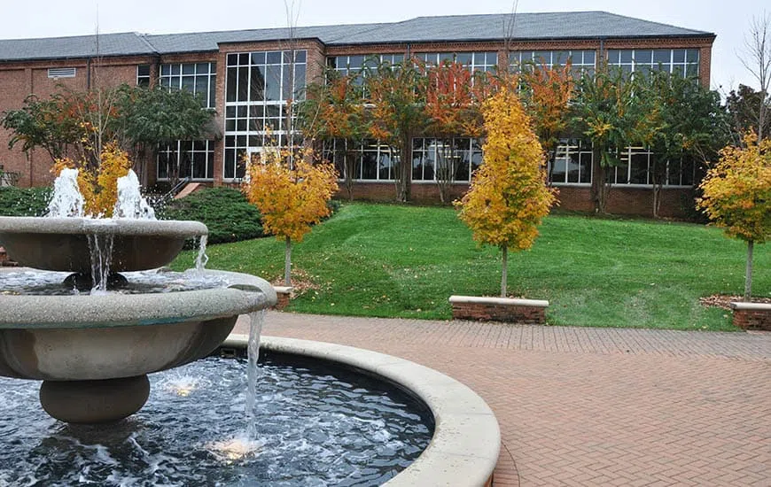 A three-tiered fountain appears in the foreground surrounded by a brick patio. A glass-walled three story building appears in the background beyond a grassy field.