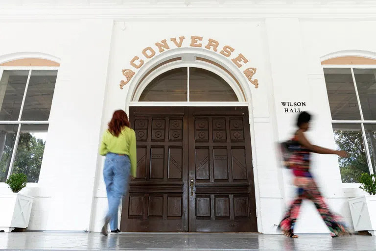 Two female students walk near wooden double doors topped by an arched window. Gold words above the arch read: CONVERSE.