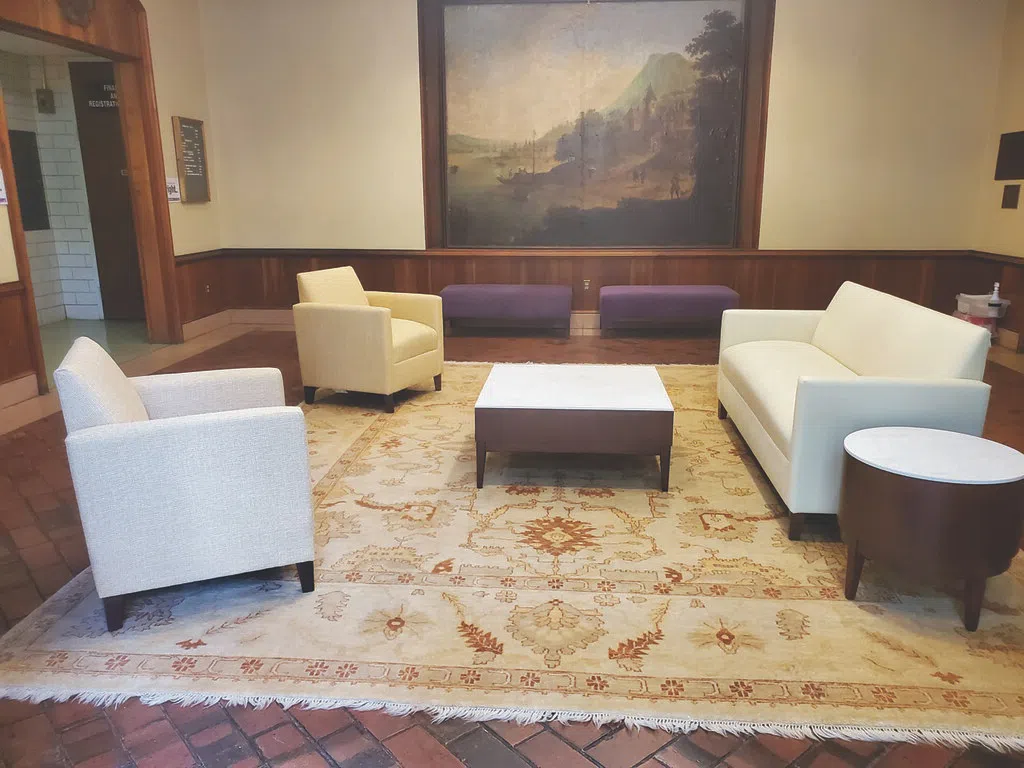 White and yellow arm chairs are positioned facing a white sofa on a large oriental rug. An oil painting of a landscape scene hangs on the wall behind the sitting area above two purple benches.