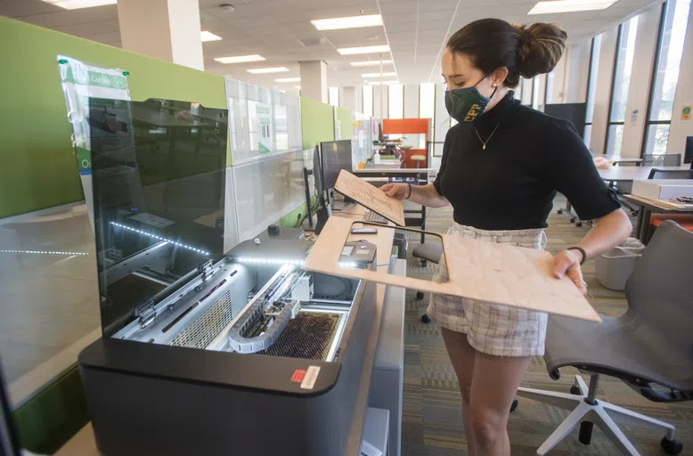 A female student standing in front of a laser cutter holding a piece of wood