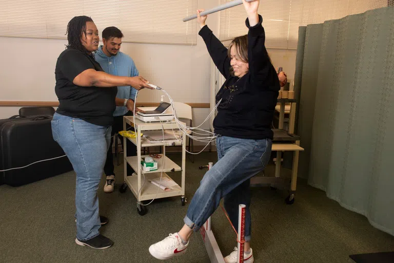 a student holds a bar above their head and balances on one foot while hooked up to a machine