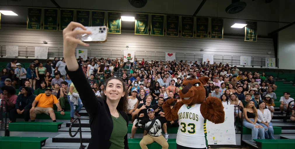 A student takes a selfie with Billy Bronco and students in the stands of a gym