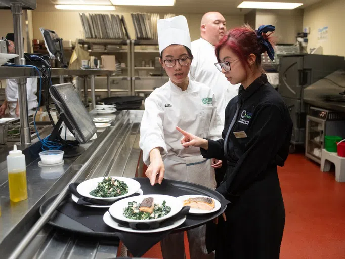 A server holds a tray of plates while a chef points to a plate