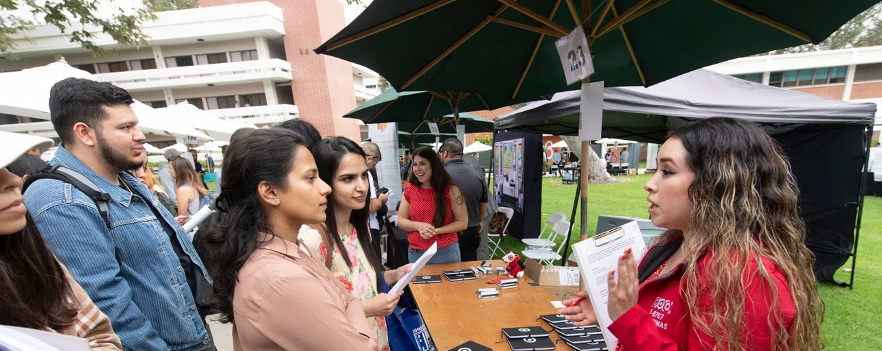 a group of students standing in front of a table listening to a representative from a company