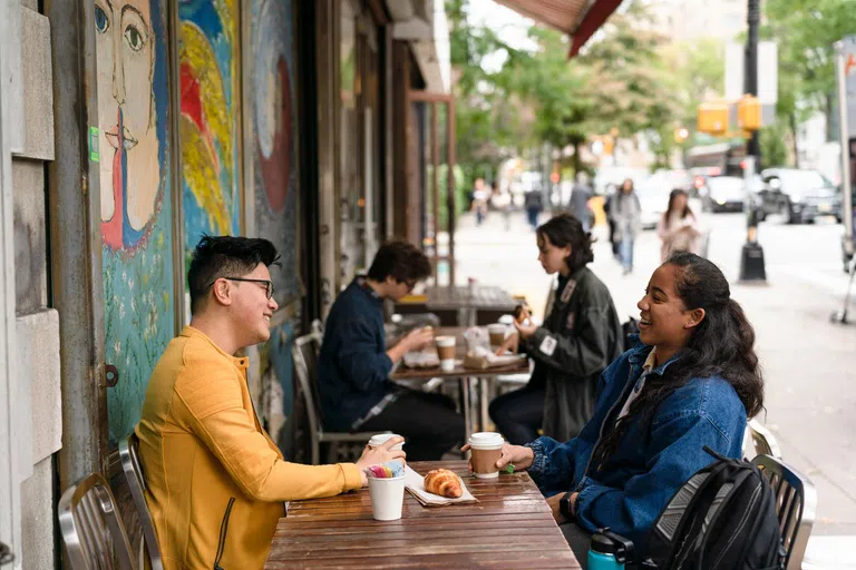 Two students, one male and one female, sit across from each other at a small wooden table. They are an an outdoor cafe and talk over coffee and pastries.