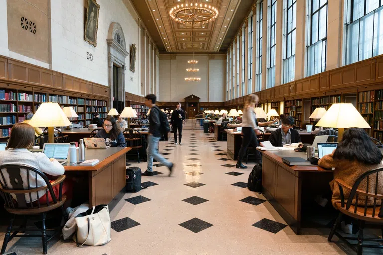 Well-lit busy study room in a library. Students sit at long wooden tables studying while people flow in and out of the rooms.