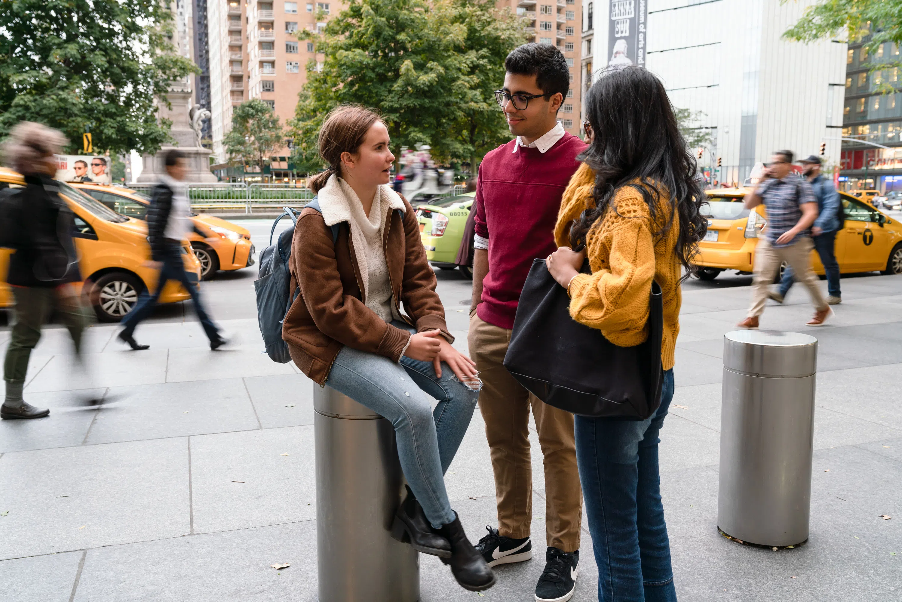 Three students sit at a busy traffic roundabout, there are blurred cars and people in motion behind them.