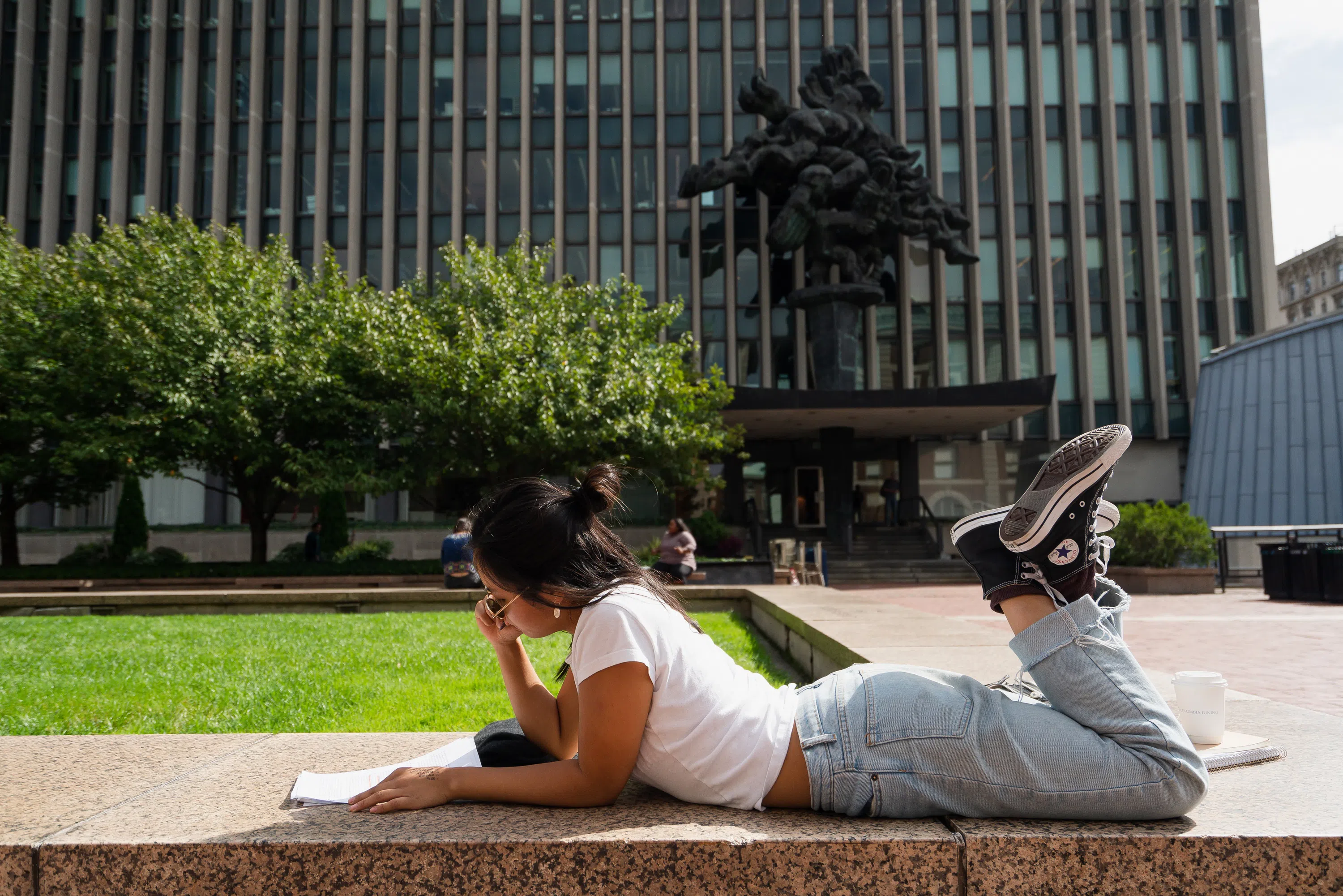 Student lounges while reading a book on a stone ledge. Behind her there are trees, sculpture art, and a glass building.