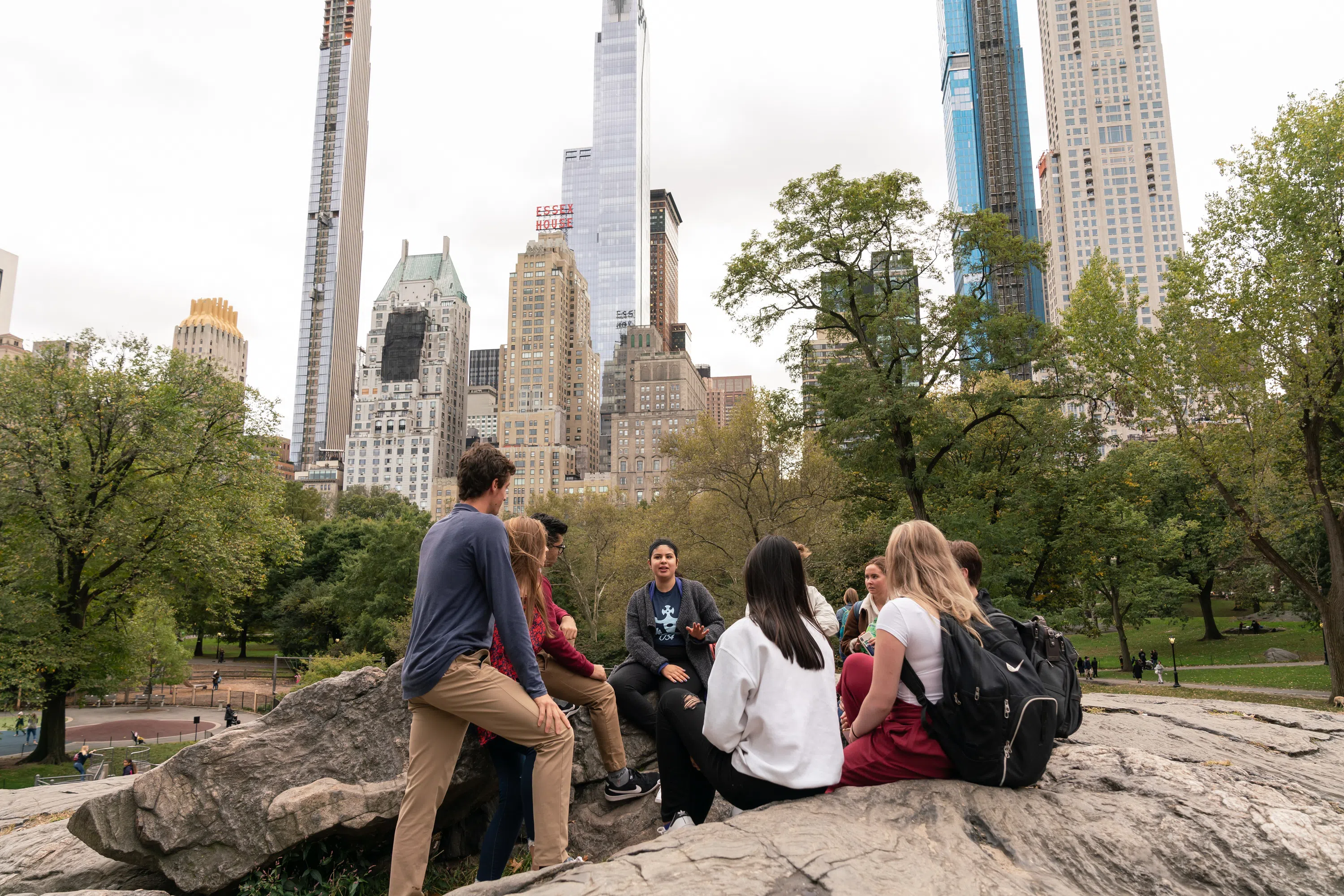 A group of students sit in a circle in Central Park, they are surrounded by greenery, rocks, and trees. There are several tall skyscrapers in the background.