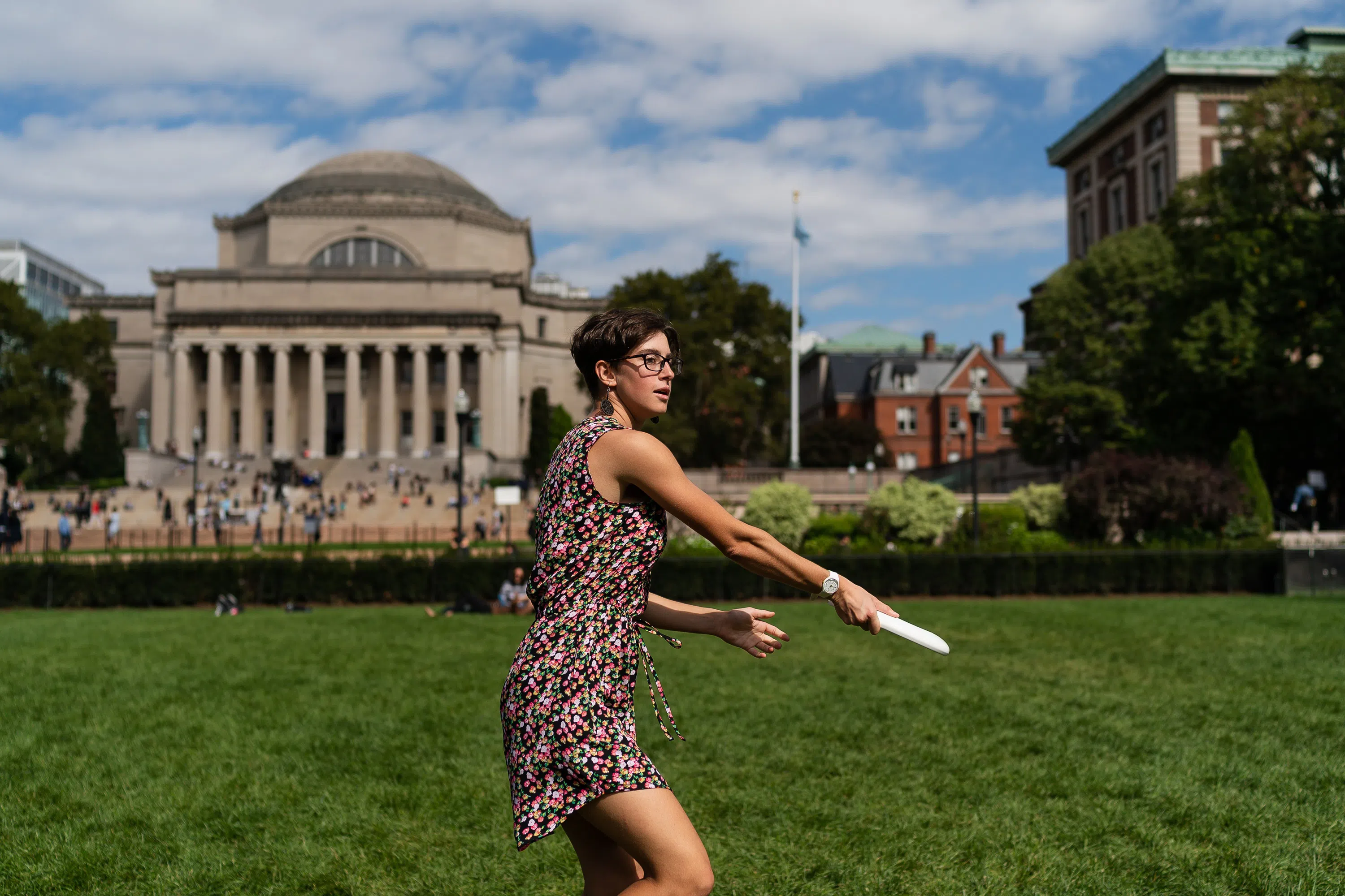 A student stands on a green lawn and throws a frisbee to a friend in the distance.