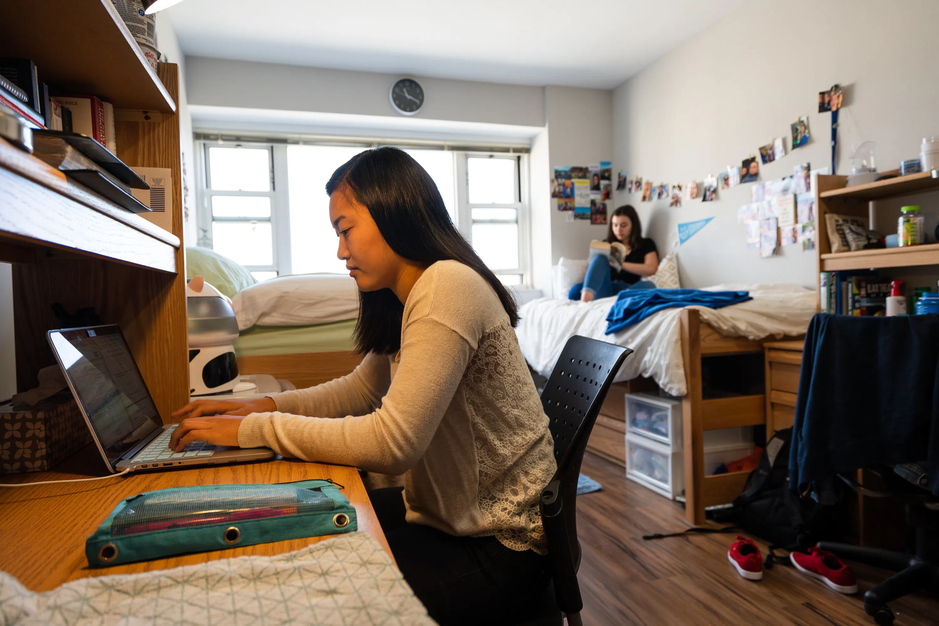 Two female students sit in a dorm room, one sits at her desk while the other is on the bed, both are studying.