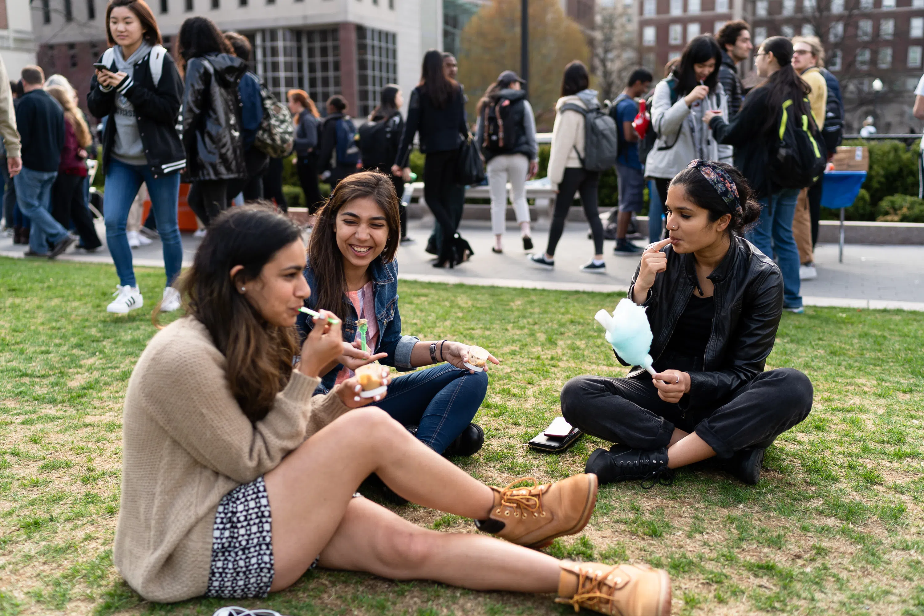 Three female students sit on the the grass in a circle while eating cotton candy and ice cream desserts.