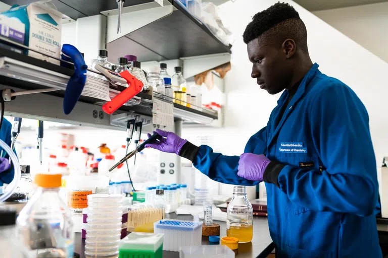 Young male student works are a lab bench, wearing a blue lab coat and pipetting materials.